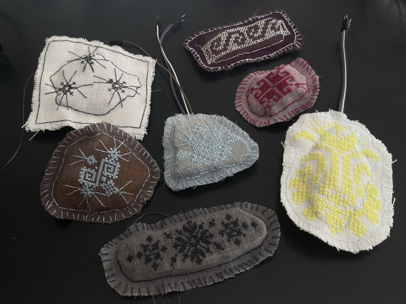 Seven stones wrapped in hand-embroidered textiles, wires leading out of each